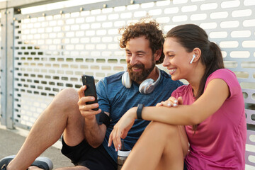 Two friends relax on the pavement after a jog, smiling as they review fitness updates on their phones, embodying a healthy lifestyle in an urban area.