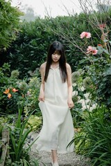 Young woman in a flowing white dress walking through a lush garden filled with flowers.