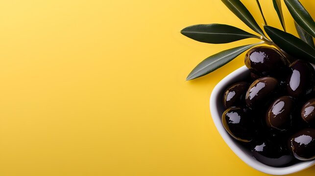 Black olives in white ceramic bowl with green leaves on bright yellow background, minimalist food composition with copy space for text or design.