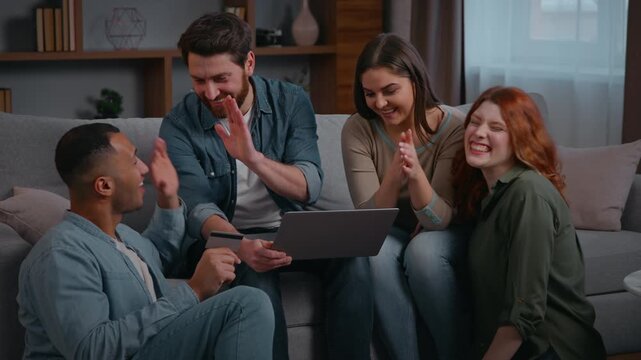 Joyful happy multiracial four friends shopping laptop celebrating achieve applauding yes gesture successful winning money online bet casino at home party on couch diverse men women using credit card