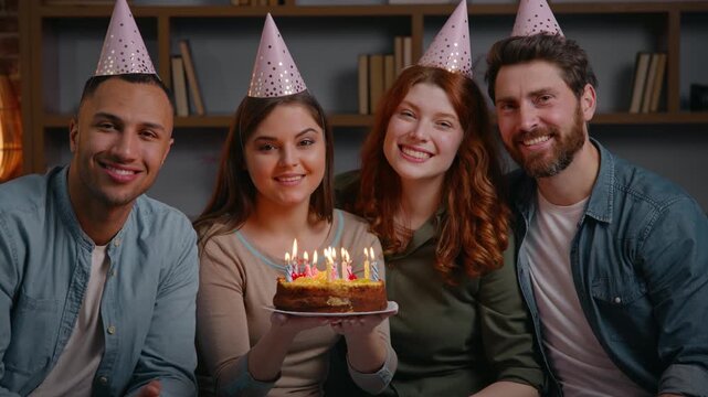 Four multiracial friends in party caps celebrate birthday holding cake smiling looking at camera portrait festive event at home diverse buddies carefree faces together posing indoors people men women