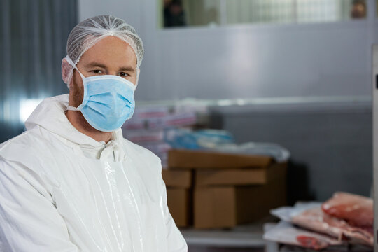 Standing man wearing protective suit and mask in processing plant, with meat, boxes, copy space