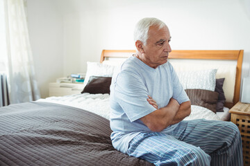 White-haired man sitting on bed edge near nightstand with books, pillows, gazing right in bedroom