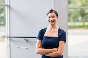Smiling woman in twenties standing in lobby near large glass window pane and frame, arms crossed