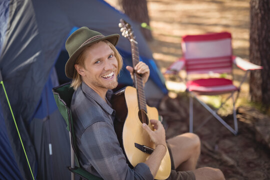 Man playing acoustic guitar in green chair at campsite with tent and striped red-and-white chair - Powered by Adobe