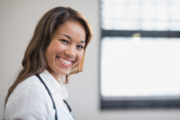 Mid adult Asian doctor wearing stethoscope standing in medical office smiling at camera, copy space