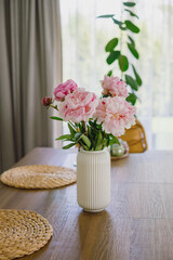 Blooming pink peonies in a white vase on a wooden dining table in a modern minimalistic home interior