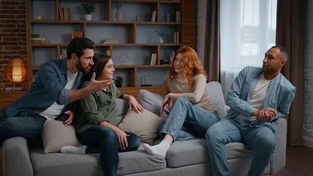 Multiracial four friends at home party talking on couch emotional discussion arguing smiling diverse people communicate in living room women girls men guys together conversation debate problem discuss