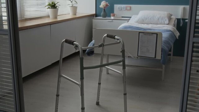 Tilt down shot of hospital ward showing metal walker in foreground, single bed with medical form attached to it, bedside table and potted plants on windowsill in background