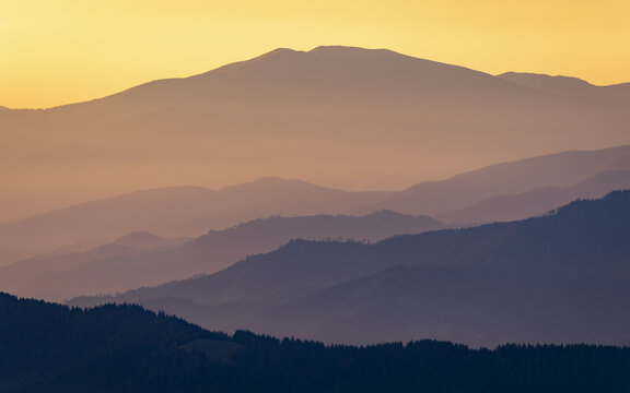 View of dusky mountain silhouettes cascading into the horizon under a gradient sky of warm yellows and oranges, Styria, Austria. - Powered by Adobe