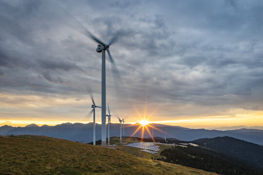 View of wind turbines spinning against a dramatic sunset sky, contrasting with the rugged mountain terrain, creating a powerful scene, Lachtal, Styria, Austria. - Powered by Adobe