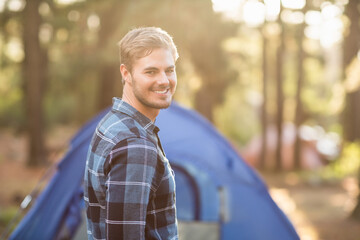 Smiling man standing in sunlit forest clearing, with blue camping tent pitched behind him