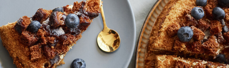 Website header shot of slice of cake topped with blueberries and chocolate chunks on plate with golden spoon beside, whole cake partially visible on side, no people present