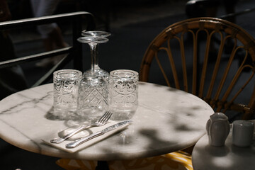 Elegant close-up of a marble café table set for two, featuring vintage-style glassware, cutlery on a napkin, and warm natural light casting soft shadows. Romantic, Parisian or Mediterranean vibe