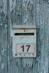 Old metal mailbox with number 17 on a weathered wooden wall with peeling blue paint. Rustic architecture, retro detail, textured surface.