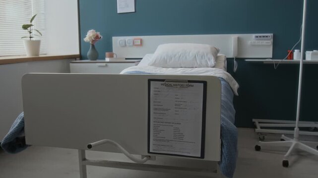 No people view of medical history form attached to footboard of patients neatly made hospital bed with bedside table and IV stand against blue wall