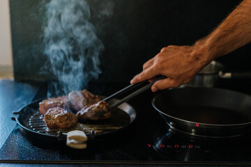 Close-up of a man’s hand using tongs to cook juicy pieces of meat on a hot grill pan with smoke rising in the kitchen. Home cooking and delicious food preparation scene.	