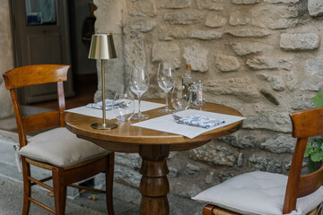 Outdoor restaurant table with elegant table setting, wine glasses, and brass lamp near old stone wall. European cafe atmosphere.