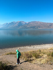 Fototapeta premium Child in green jacket stands at Jackson Lake shore with Grand Teton mountains under a clear blue sky.