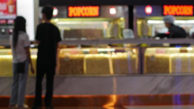 Blurred cinema concession stand with customers buying popcorn and drinks under colorful neon lighting at movie theater