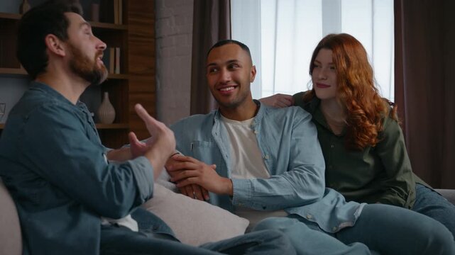 Three multiracial friends at home party talking Caucasian man sharing news Latino African American guy listening Caucasian woman multiethnic buddies diversity people men friendly spending time smile
