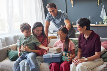 Fototapeta premium Family group including middle aged Caucasian woman and man, young adult woman, young boy and girl sitting on sofa giving gifts