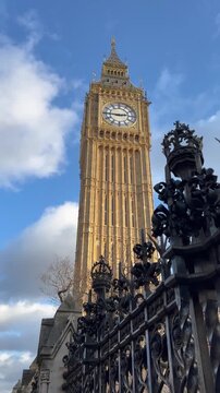 Low-angle of London's Big Ben clock tower, known as the Elizabeth Tower. Grandeur of ornate Gothic architecture and famous clock face, with foreground element of decorative wrought-iron fence.