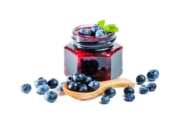 Jar of blueberry jam with berries, leaves and wooden spoon isolated on white background.