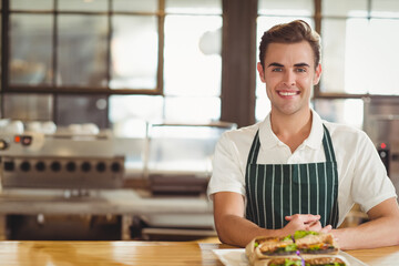 Smiling man leaning on wooden counter in bright café, with white tray of sandwiches, copy space
