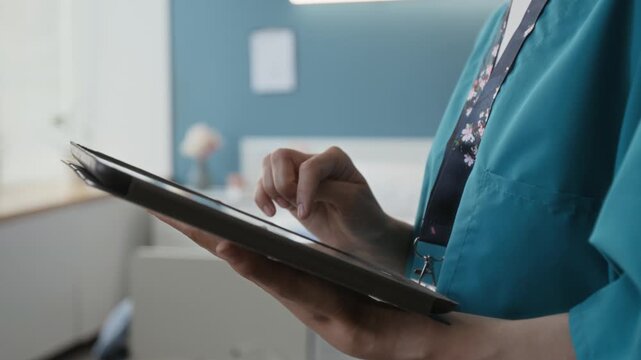Close-up tilt up shot of concentrated Caucasian young nurse wearing scrubs with badge using digital tablet in hospital ward