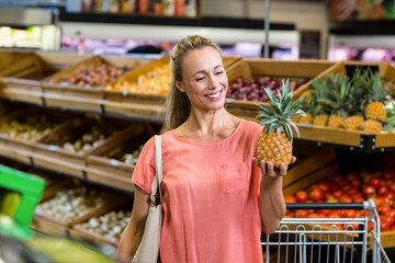 Mid adult woman holding pineapple while smiling in supermarket produce aisle, with grocery cart