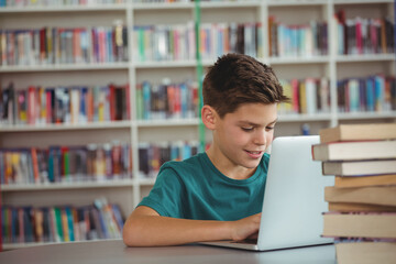 Child typing on silver laptop at library study table, with stack of hardcover books
