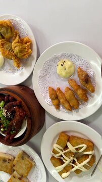 Top view vertical footage of assorted dim sum dishes including har gow dumplings, turnip cake, braised chicken feet, fried wontons, and crispy dumplings served on white plates and bamboo steamers.
