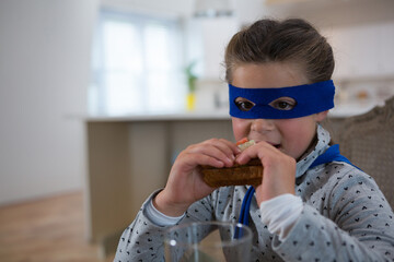 Biting girl child wearing blue mask and cape sitting at kitchen table, with sandwich, copy space