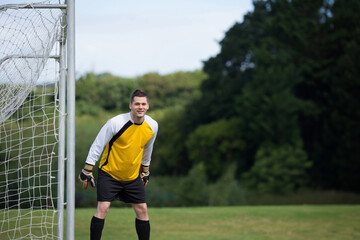 Male goalkeeper standing by goal with netting on pitch wearing yellow jersey, gloves, copy space