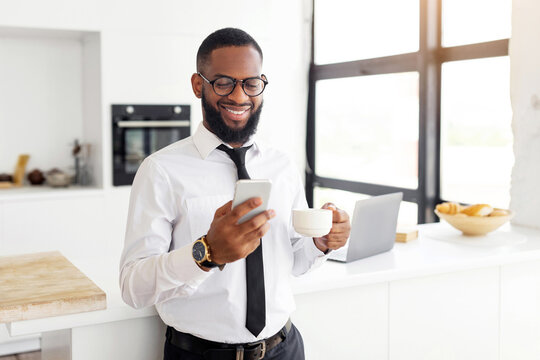 A man in a formal shirt and tie stands in a bright kitchen, smiling as he looks at his smartphone. He holds a coffee cup in one hand and is beside a laptop and a bowl of pastries.