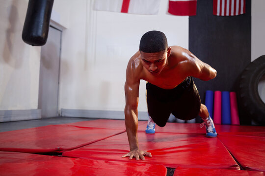Performing man holding one-armed push-up on red padded mat in gym training room, with punching bag