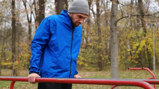 Man in blue sports jacket exercising on parallel bars in autumn park, training arms and upper body strength. 4K horizontal footage.