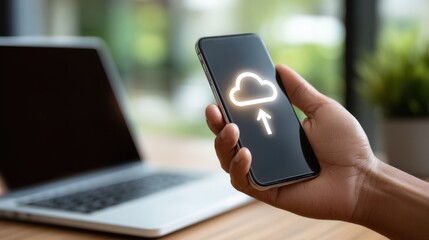 A hand holding a smartphone with a cloud upload icon, signifying data transfer or storage, in a modern workspace.