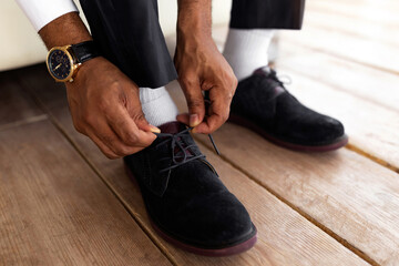 A man in formal attire sits on a wooden floor, meticulously tying his black suede shoes. He wears a...