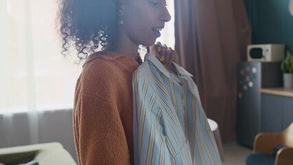 Young Biracial woman selecting between two outfits on wooden hangers in bright, sunlit apartment, reflecting style and daily routine - Powered by Adobe