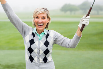 Senior woman wearing argyle sweater raising arms and celebrating shot on golf fairway with club