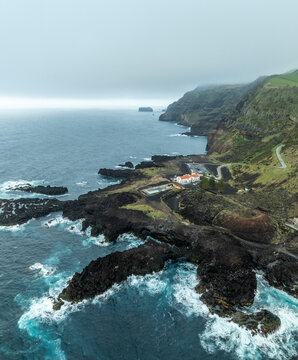 Aerial view of the rugged coastline meeting the turbulent sea, the stark black lava rocks contrasting with the white foam of crashing waves near Termas da Ferraria, Ferraria, Azores, Portugal.