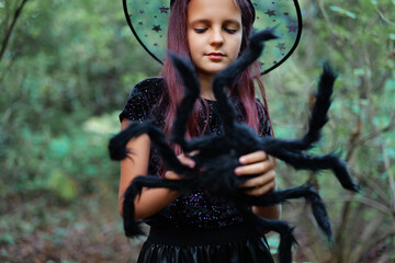 Naklejka premium Young girl in witch costume holds a large spider in a forest setting during Halloween celebrations