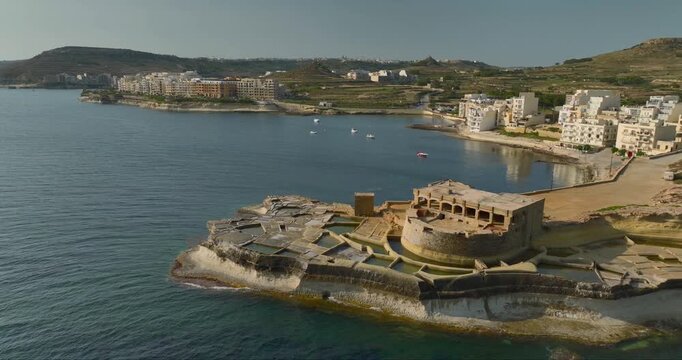 Aerial view of the stark, stone Qolla l-Bajda Battery standing guard over the tranquil, turquoise waters of the bay, casting long shadows, Gozo, Malta.