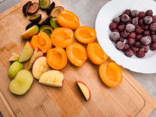 Sliced apples, apricots, and plums resting on wooden cutting board with frozen cherries for compote