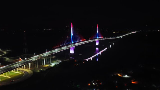 Slow aerial dolly in over illuminated Puente H&eacute;roes del Chaco at night, lit in red, white, and blue with reflections on the Paraguay River.
