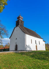 Naklejka premium A medieval Romanesque church with a distinctive white exterior and wooden shingled roof stands on a grassy hill under a dramatic and colorful sky with dark, cloudy formations.