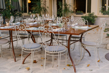 Charming outdoor restaurant terrace with elegant wrought iron chairs, wooden tables, and glassware set for dinner. Cozy European courtyard dining atmosphere, perfect for summer evenings.