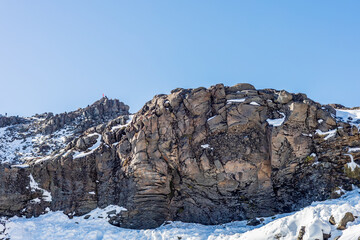 A rugged mountain cliff with textured rock formations lightly covered in snow, standing against a clear blue sky